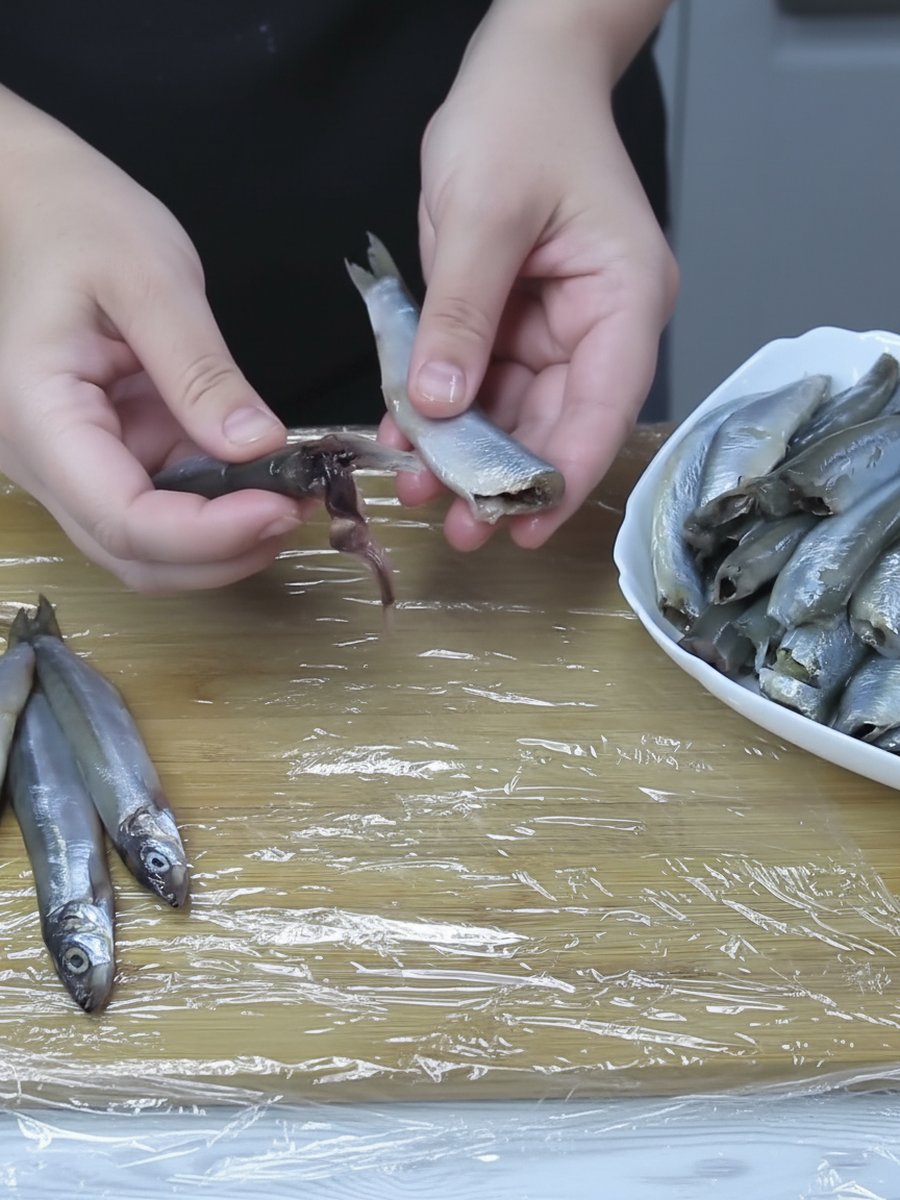 Руки потрошат мойву, рядом целая рыба и миска с очищенной мойвой